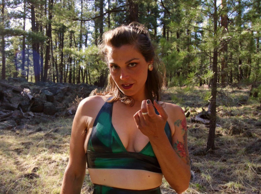 Dominatrix Mistress Pomf stands in the forest, cloaked in shiny green latex and as she beckons you closer with her finger.