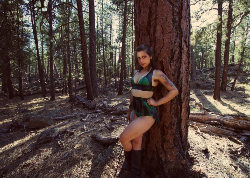 Dominatrix Mistress Pomf stands amidst a forest of ponderosa holding her riding crop as she deems your intent worthy or not. 