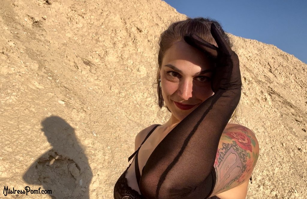 Dominatrix Mistress Pomf smiles as She looks at you from the desert. She cups Her face to block out the sun. Her arms are cloaked in sheer, black opera gloves.