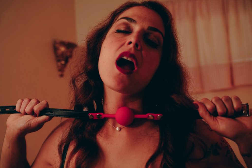 Mistress Pomf, Colorado Dominatrix, holding a black leather ball gag with a pink ball in a sensual boudoir portrait