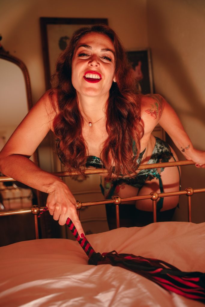 Mistress Pomf, Colorado Dominatrix, leaning over a brass bed in black and green lace lingerie and stockings while holding a red and black flogger