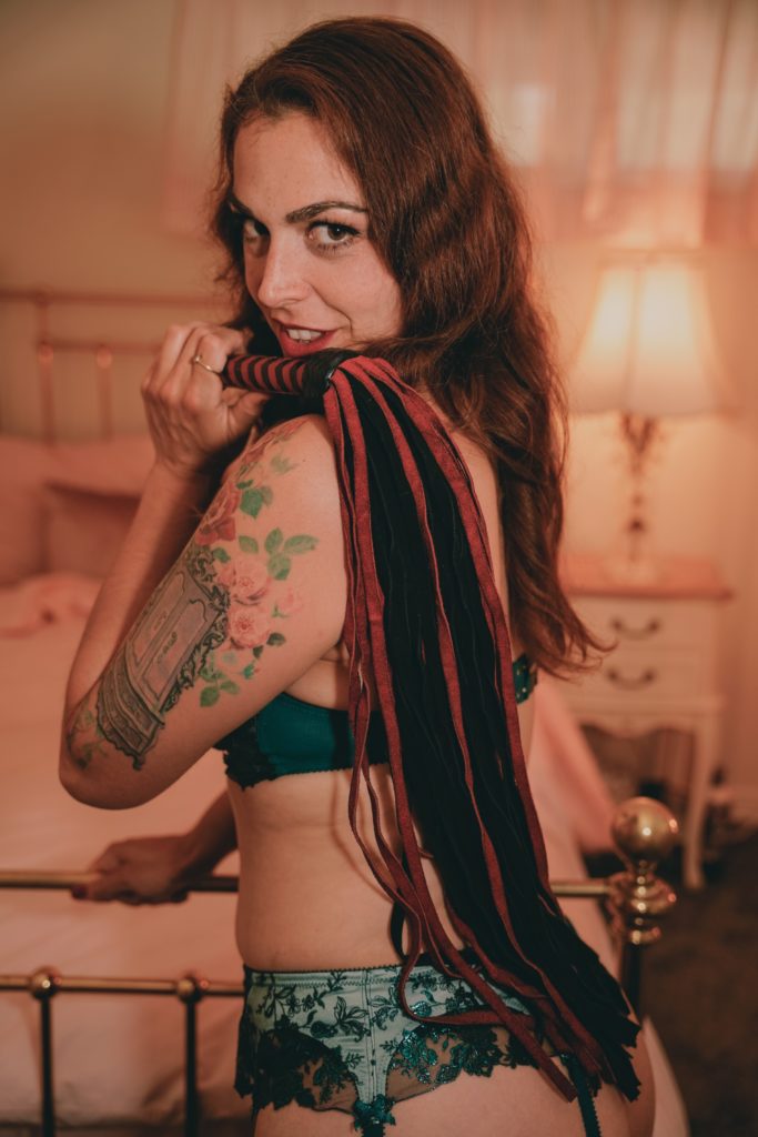 Mistress Pomf, Colorado Dominatrix, posing in black and green lace lingerie with a red and black suede flogger over her shoulder