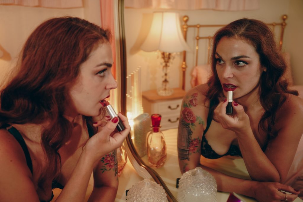 Mistress Pomf, Colorado Dominatrix, applying red lipstick while looking into a vintage bedroom mirror