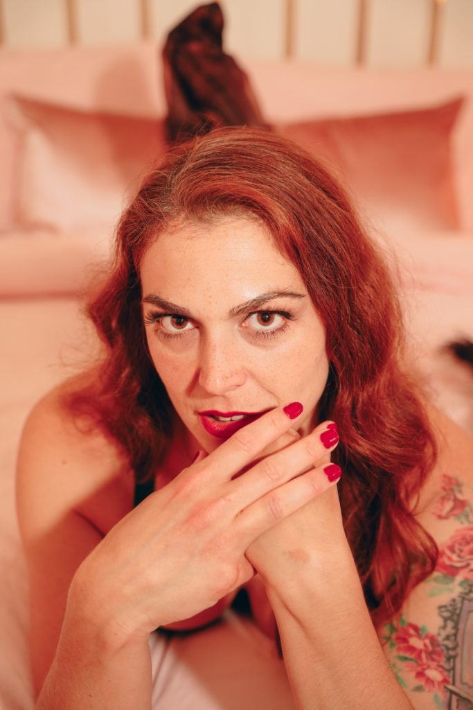 Mistress Pomf, Colorado Dominatrix, reclining on a bed with red lipstick and burgundy nails in a warmly lit boudoir portrait