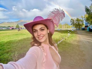 Mistress Pomf in a pink feathered cowgirl hat at an outdoor festival in Colorado, with a rainbow over the mountains